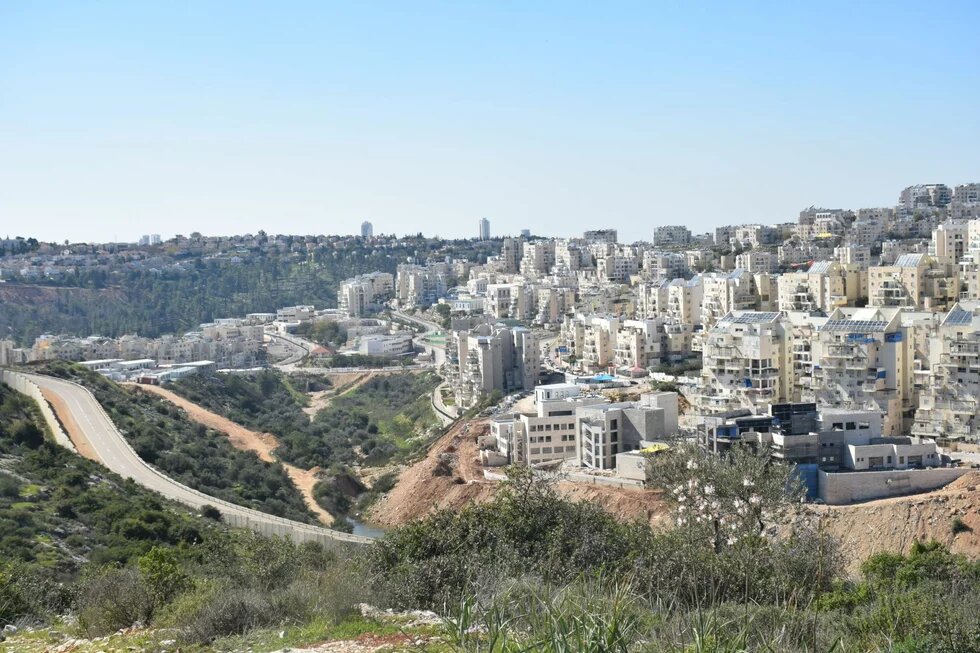 Photo: A cityscape with multiple residential buildings along a hill and a road. Green hills and trees in the background.